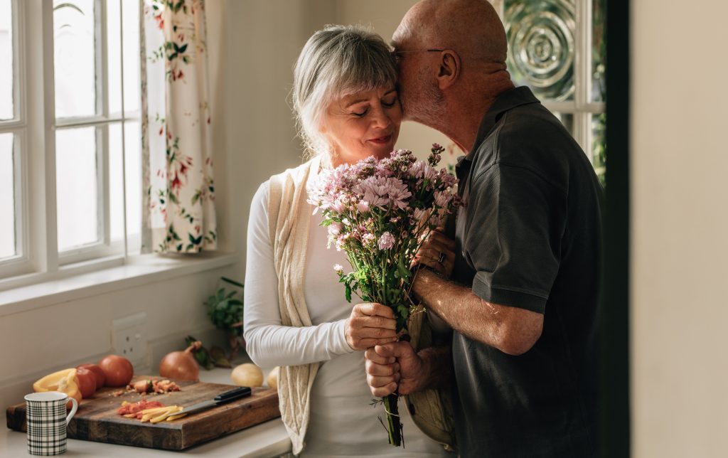 Senior couple standing in kitchen holding a bunch of flowers. Senior man kissing his wife holding her hand at home.